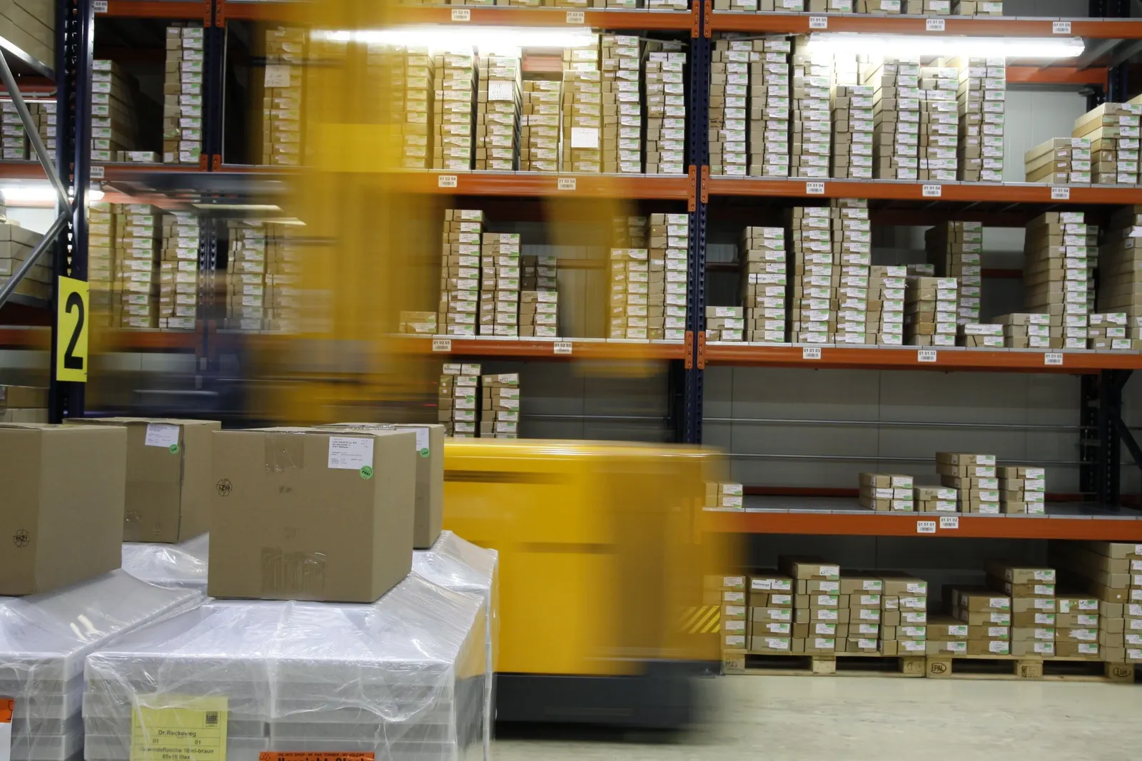 Estanterías de almacén con cajas ordenadas y etiquetas de ubicación, con una carretilla en movimiento